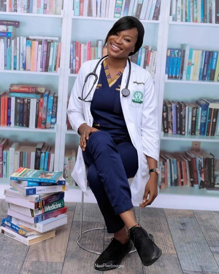 An alumna in scrubs and a white coat beside a stack of books